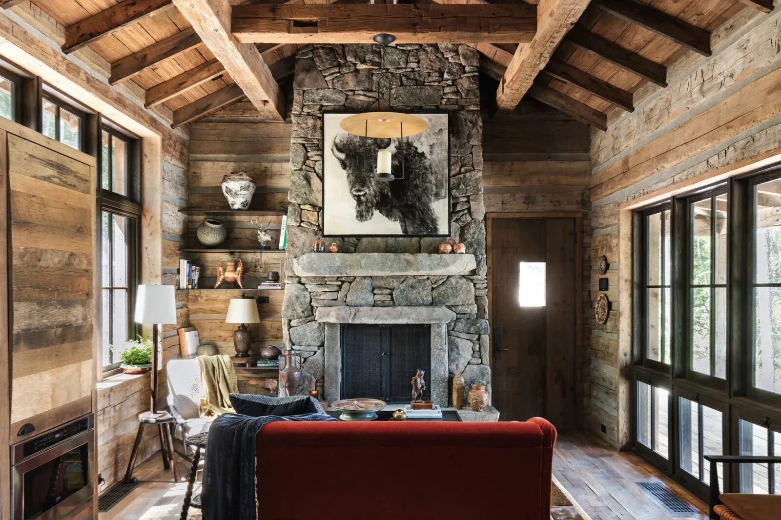 Living room of the Whitefish Cabin in Montana by Cravotta Interiors: hand-hewn timber ceiling, stone fireplace under a bison portrait, and a rust-red sofa.
